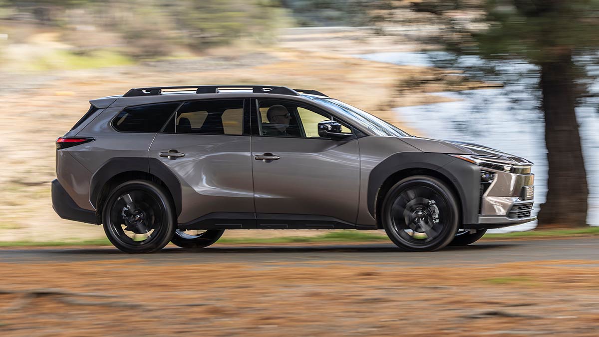 Side profile view of bronze Toyota bZ4X Touring SUV driving on treelined rural track near water crossing.