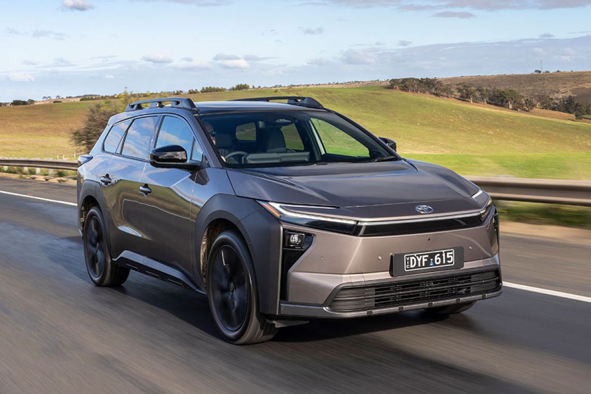 Bronze Toyota bZ4X Touring SUV driving on country highway next to undulating green paddocks and hillside.