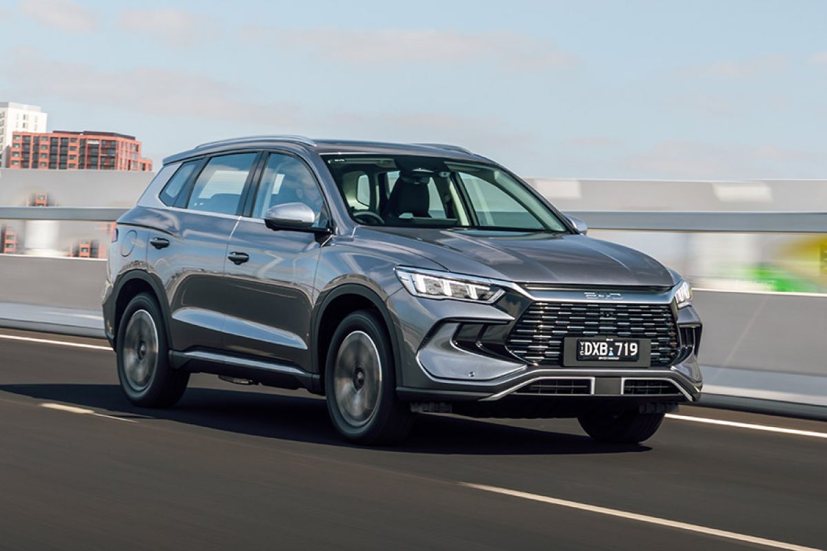 Metallic grey BYD Sealion 5 medium SUV with Victorian number plates driving on city overpass with tower blocks in background.