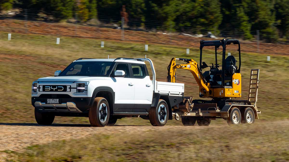White BYD Shark 6 Cab Chassis towing a trailer with excavator on a high-banked gravel road in rural area.  