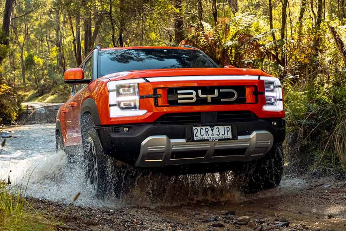 Orange BYD Shark 6 Performance ute driving through water crossing on muddy and rocky 4WD track in sunlit bushland location. 