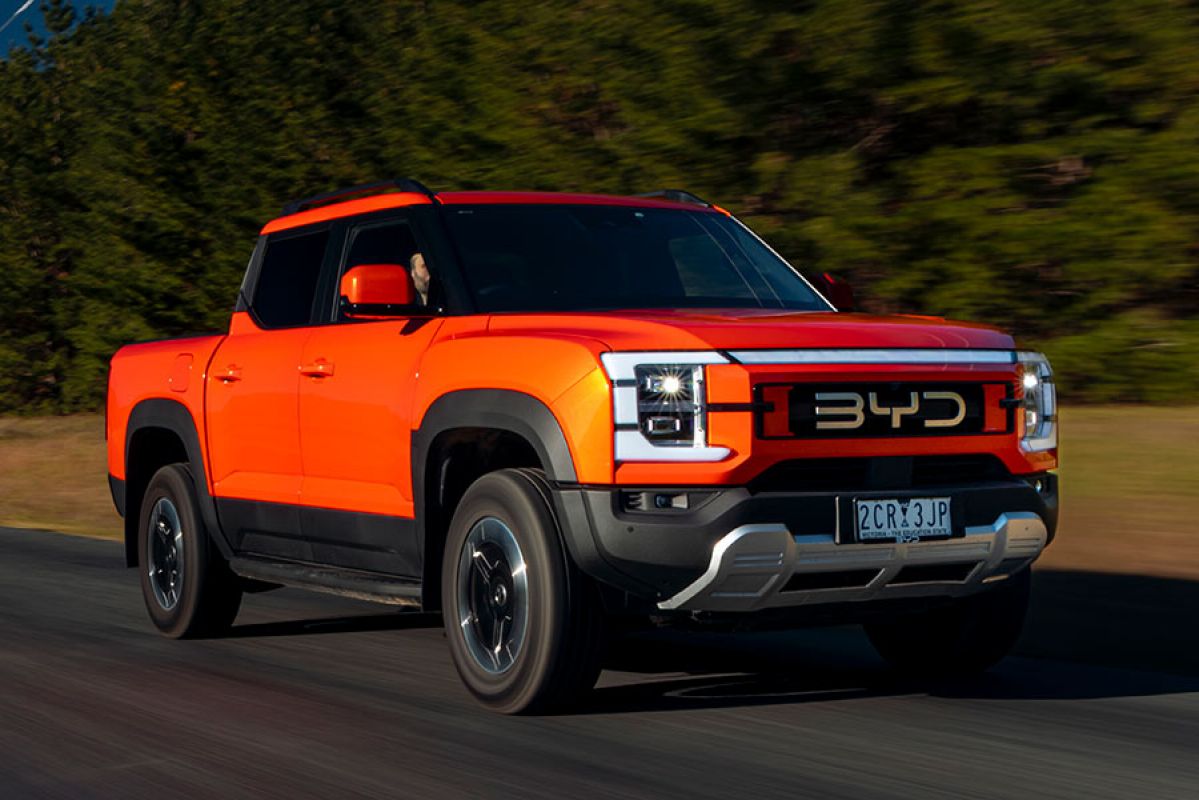 Orange BYD Shark 6 Performance ute driving on sealed country road with power lines and row of cypress trees behind.