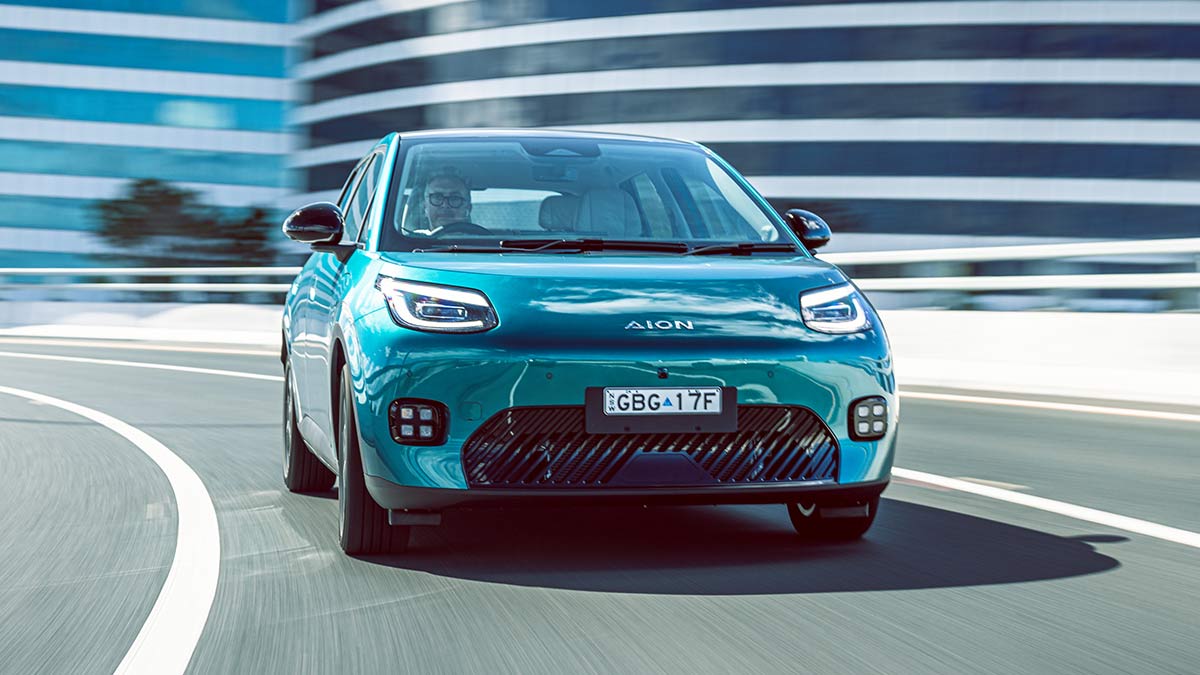 Green GAC Aion UT electric car driving on city arterial road with large white-striped buildings in background.