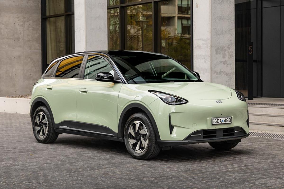 Light green Geely EX2 electric hatch with black roof, parked on grey paved area outside city building with thick concrete pillars.