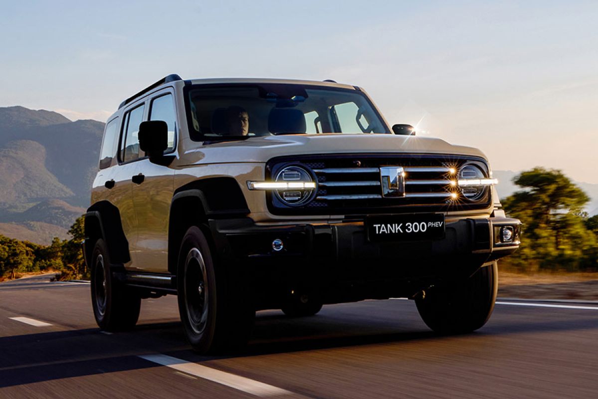 Cream-coloured GWM Tank 300 SUV driving on bitumen road in mountainous area with sunspots on 3-bar chrome grille.