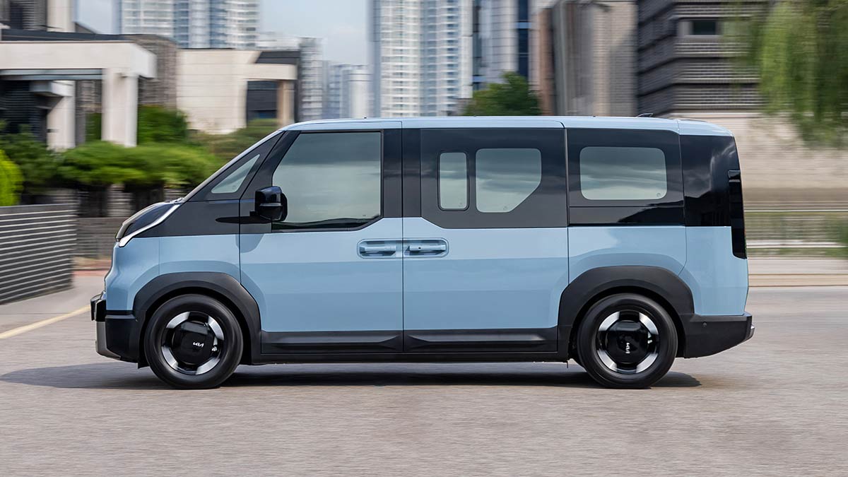 Side profile shot of blue Kia PV5 Passenger electric people-mover on road with city towers in background.