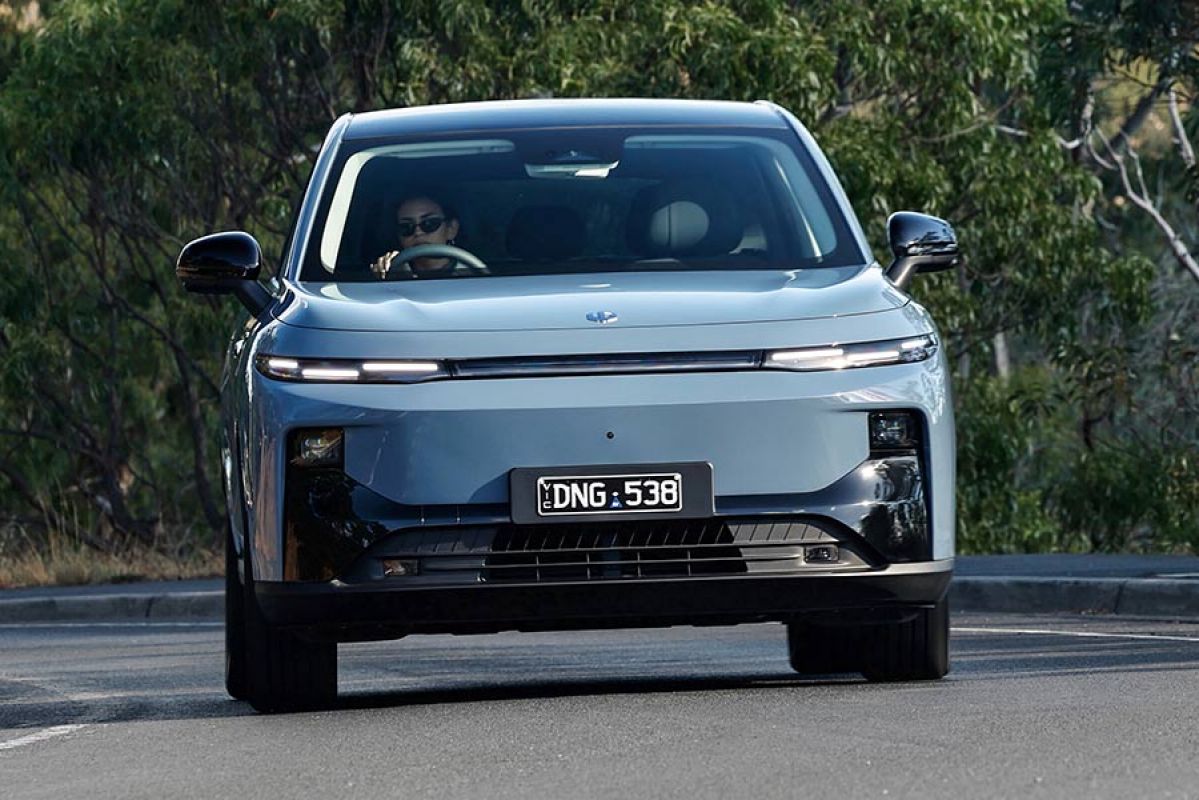 Woman with sunglasses driving blue Leapmotor B10 small electric SUV around corner on tree-lined country road.