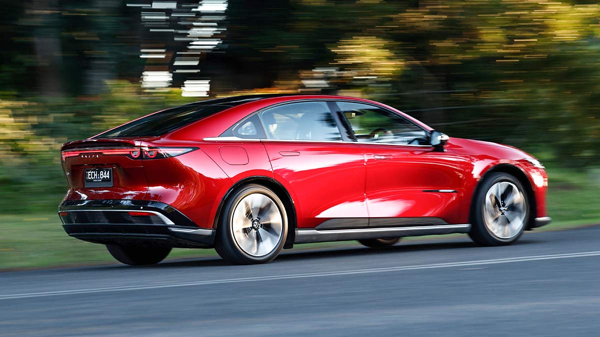Rear quarter action shot of red Mazda 6e sedan on road with blurred green and yellow sunlit background.