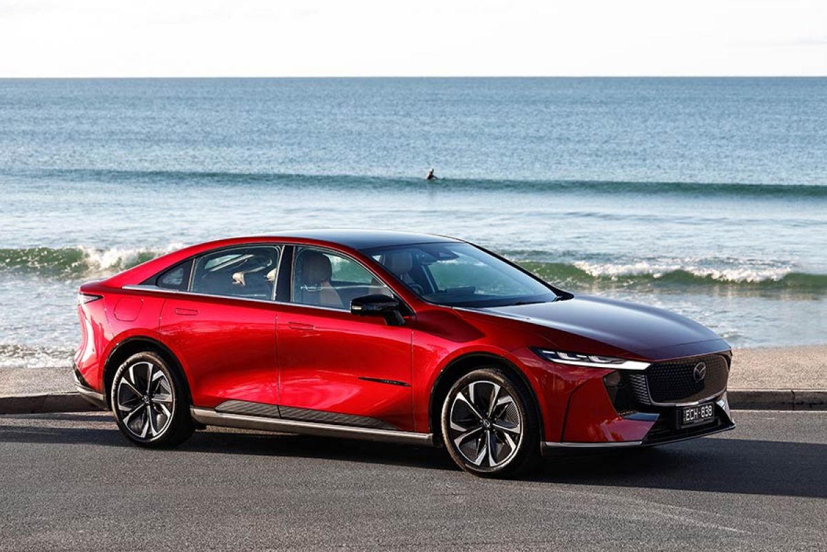 Red Mazda 6e electric sedan at beachfront carpark with lone surfer riding over wave in background.