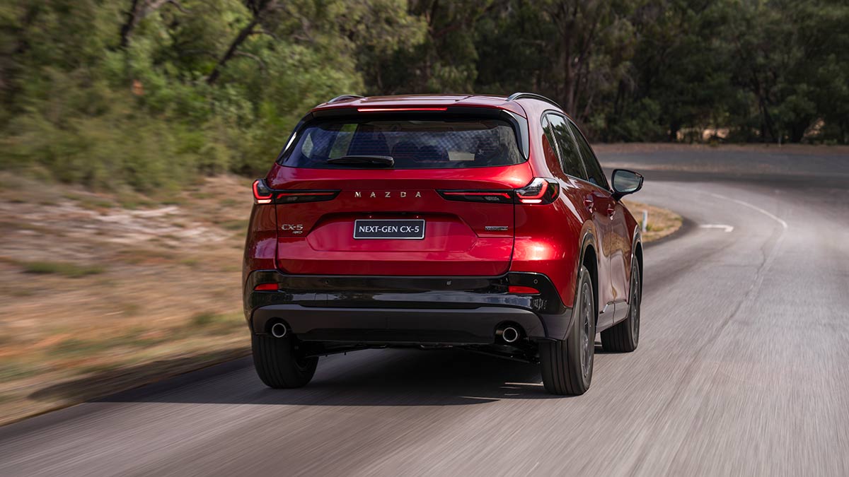 Rear view of red Mazda CX-5 SUV on narrow bitumen road approaching corner at bushy Lang Lang proving ground in Victoria.
