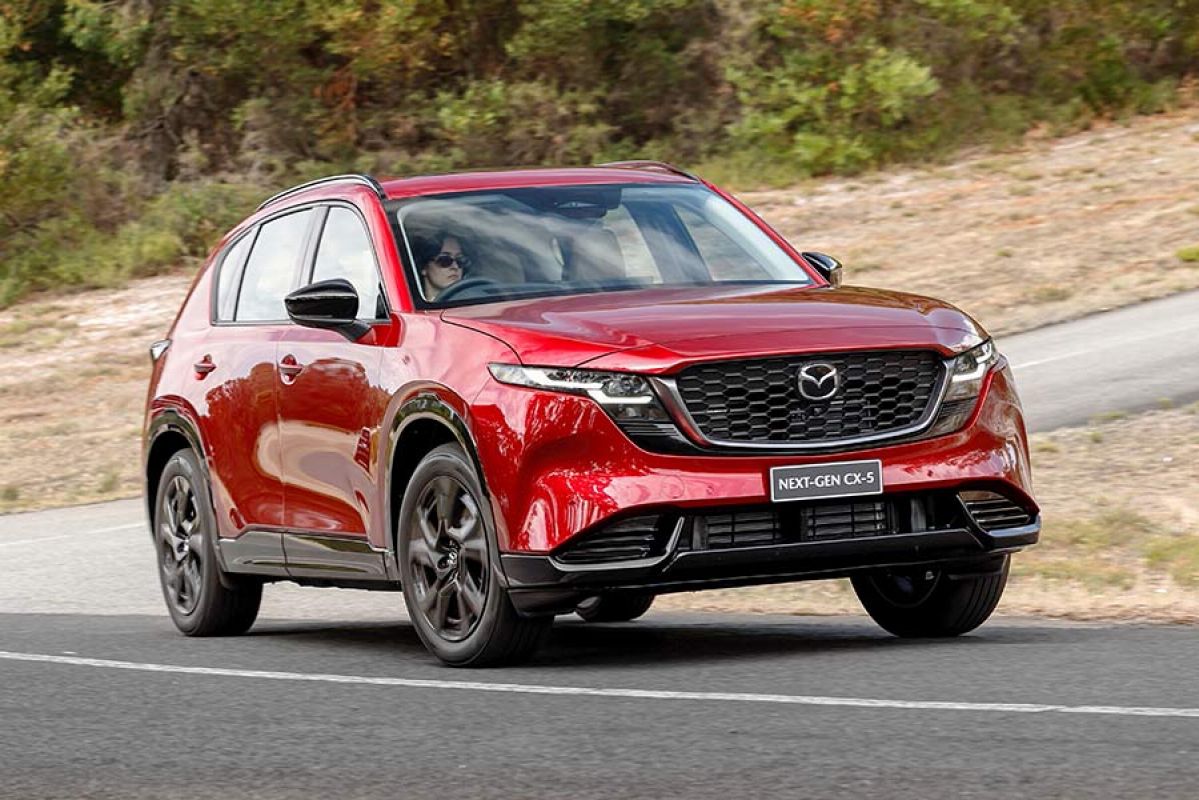 Woman with dark hair and sunglasses driving red Mazda CX-5 SUV through corner at bottom of steep hill at Lang Lang proving ground in Victoria.