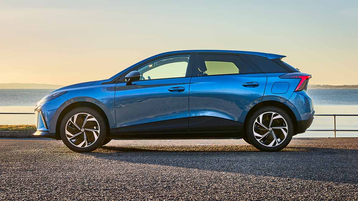 Side-on view of blue MG4 electric hatchback in seafront carpark with glassy water and orange filtered skylight in background.