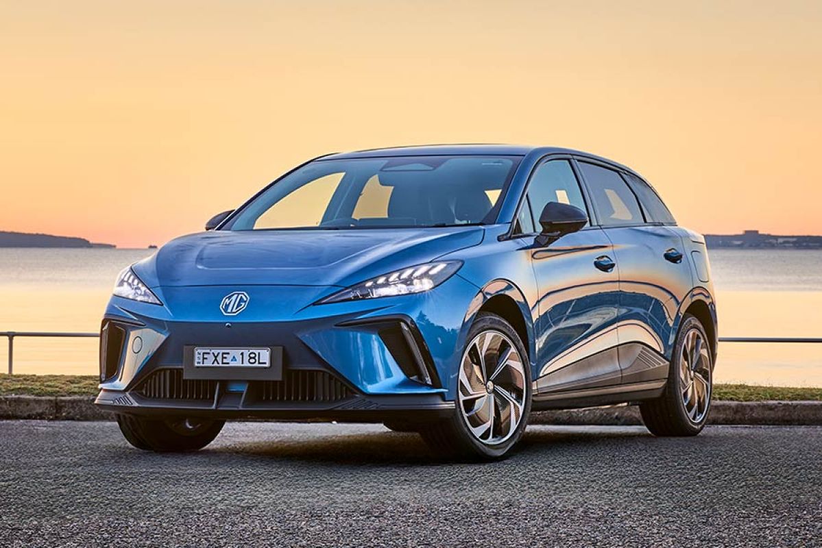 Blue MG4 electric hatchback with lights on in seafront carpark with glassy water and orange filtered skylight in background.