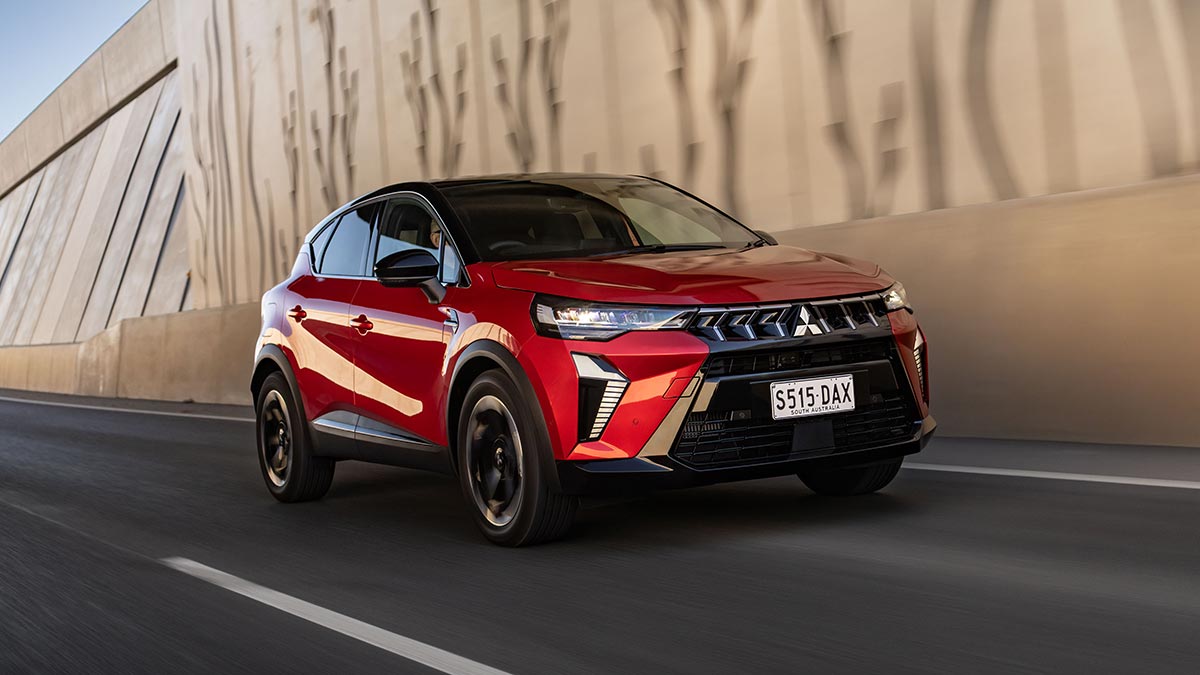 Red Mitsubishi ASX small SUV driving through high-walled underpass on multi-lane road.
