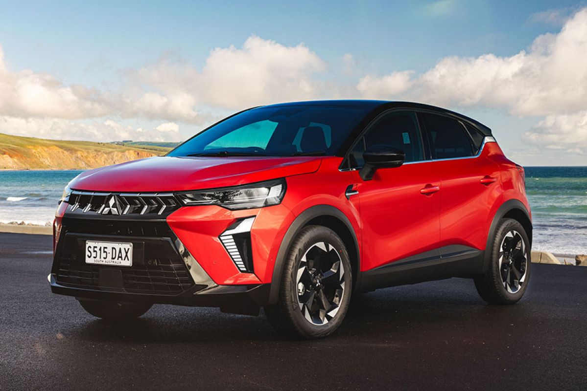 Red Mitsubishi ASX small SUV at beachfront carpark with green hills, cliffs and sea in background.