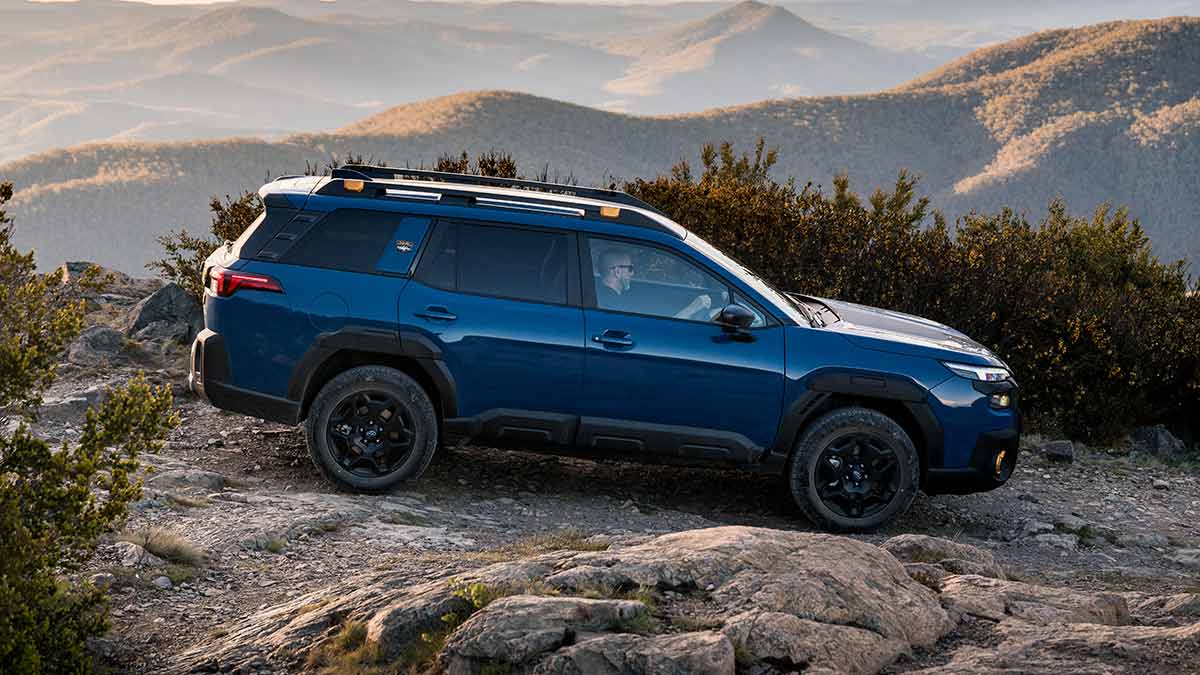 Side view of blue Subaru Outback Wilderness driving on rocky off-road trail in Australian high country with mountain ranges behind.