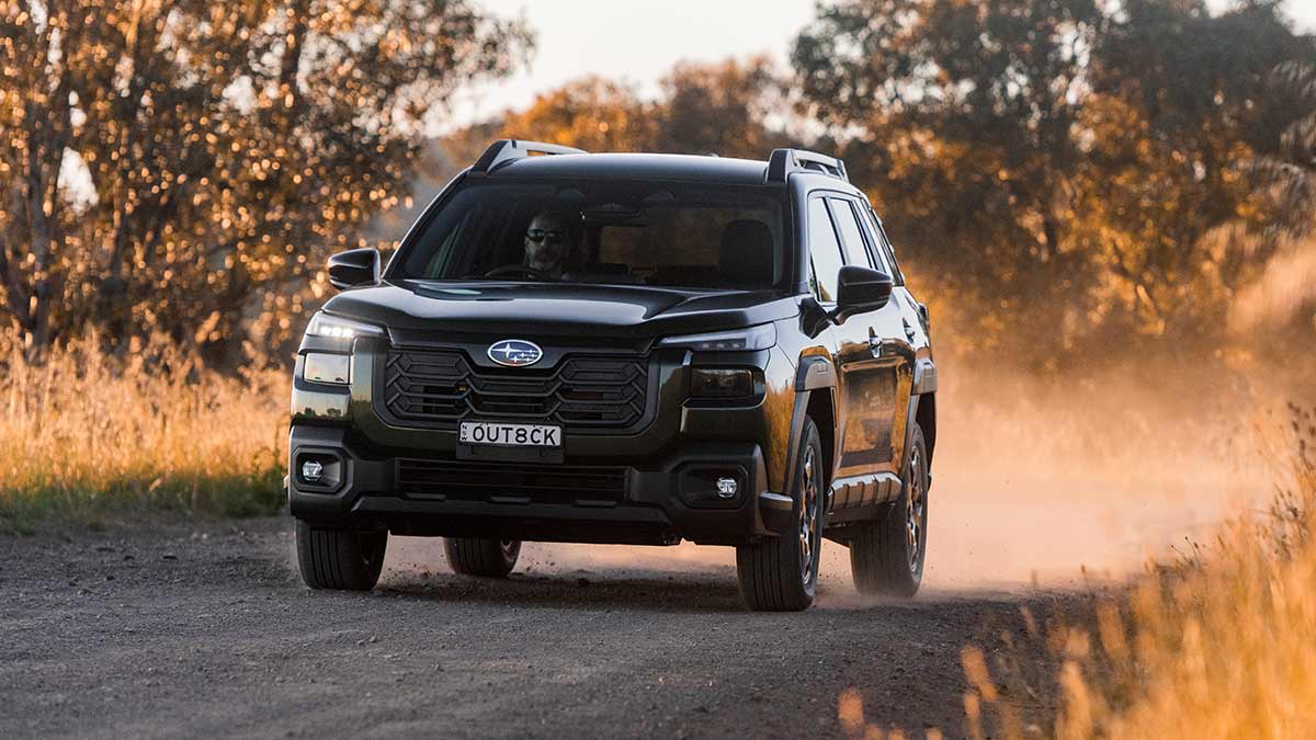 Dark Subaru Outback driving on dusty gravel road with sunlit grass and trees in foreground and background.