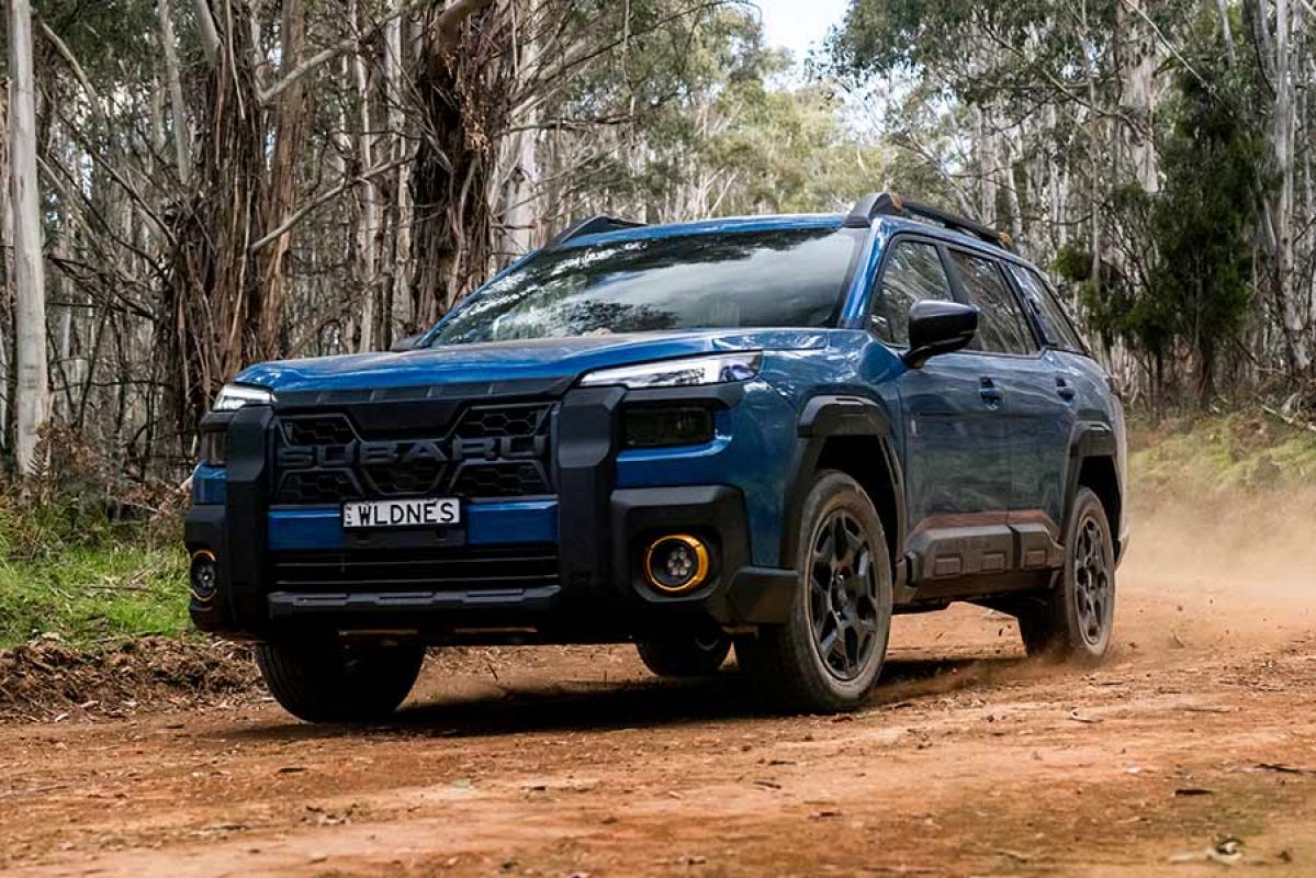 Blue Subaru Outback Wilderness SUV driving on dusty red dirt road lined with gum trees in regional Australia.