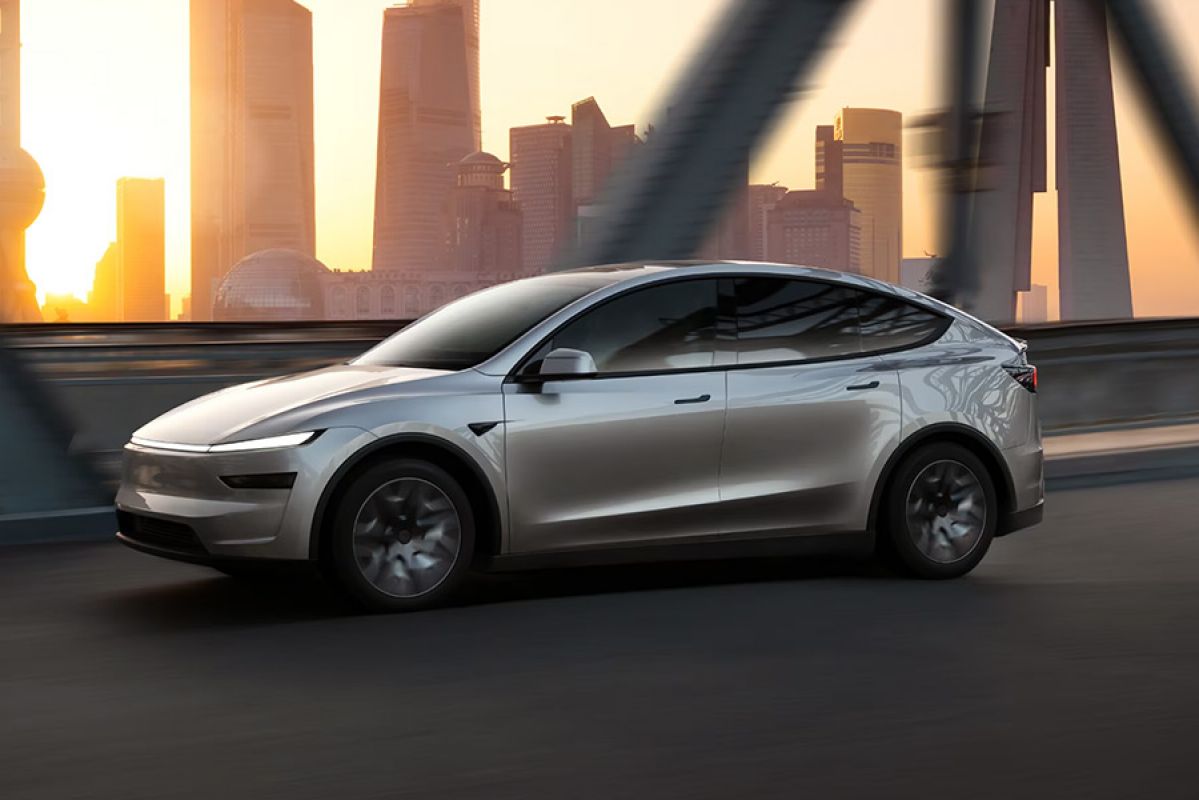 Silver Tesla Model Y L SUV driving on bridge in city with bright sunlight shining from behind towers in distance.