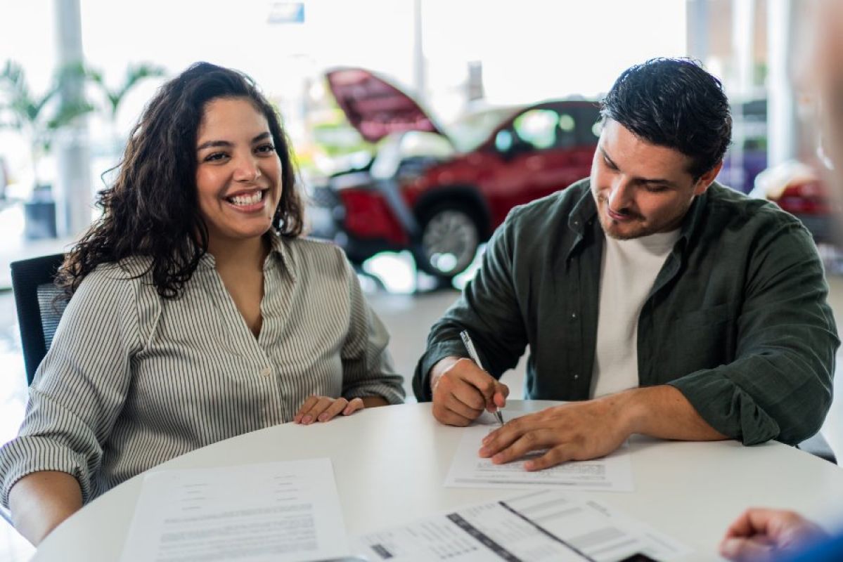 man signing finance documents while female partner smiles; lender in foreground; red car in background