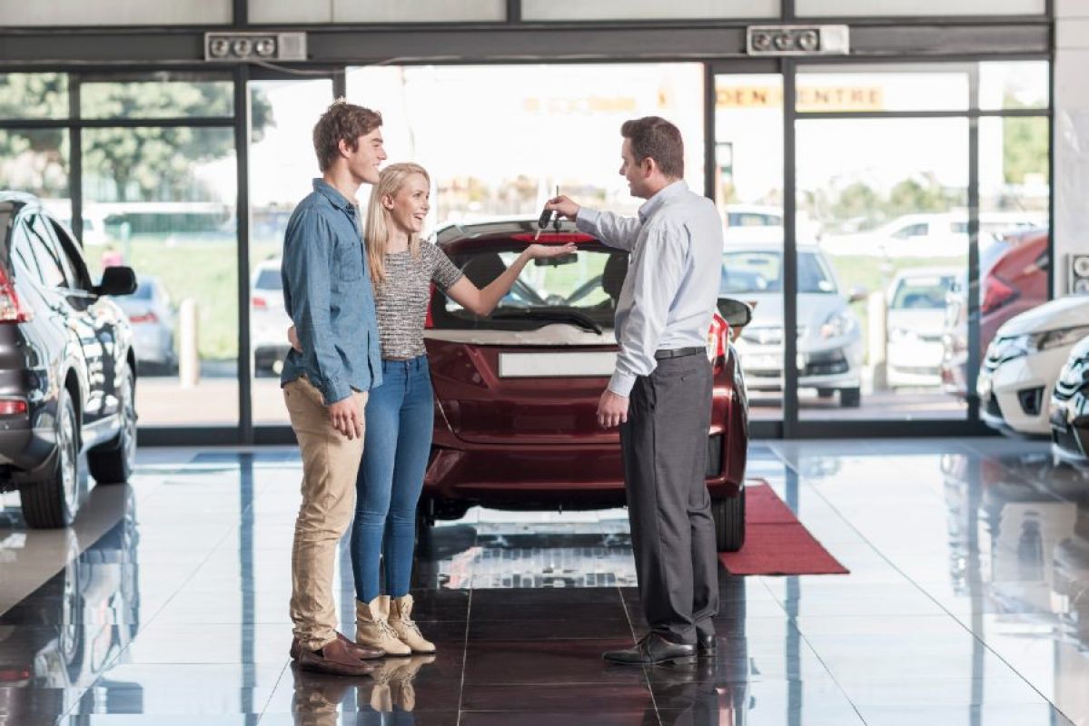 car salesman handing keys to young couple in front of small red car