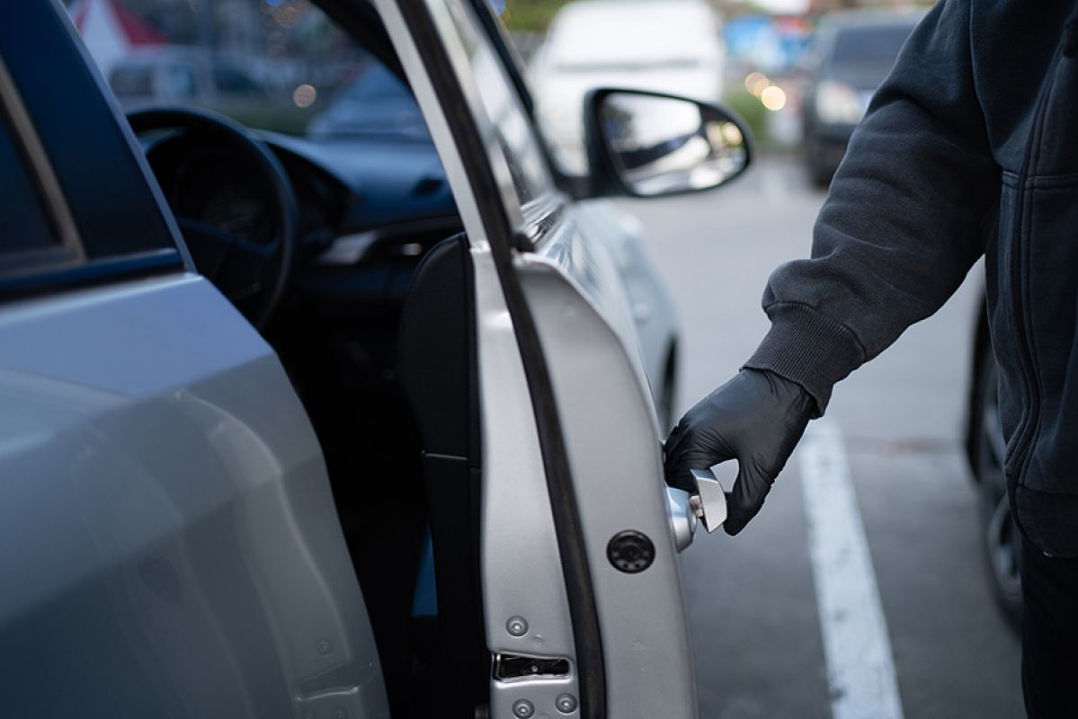 Thief wearing black jacket and black gloves opening driver’s door of silver car in carpark. 