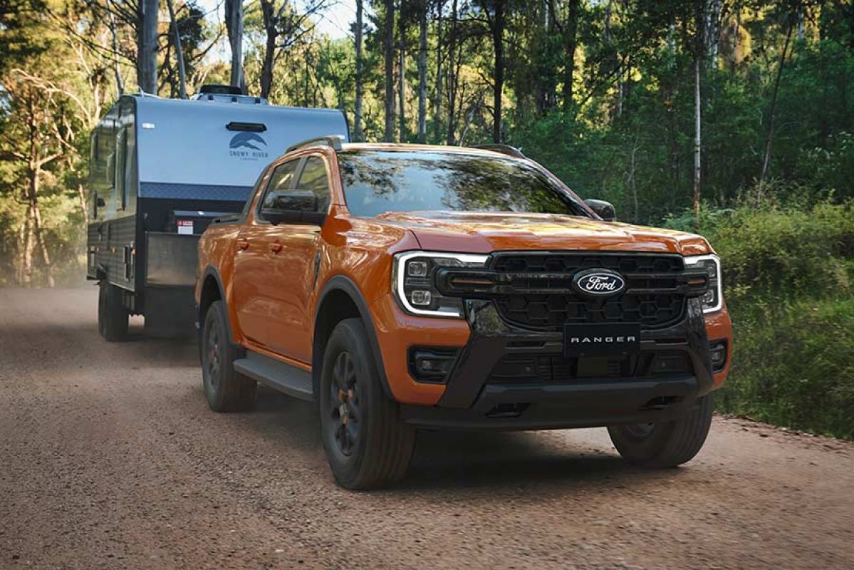 Orange Ford Ranger Wildtrak dual-cab ute towing caravan on shadowy treelined scrubby gravel road.