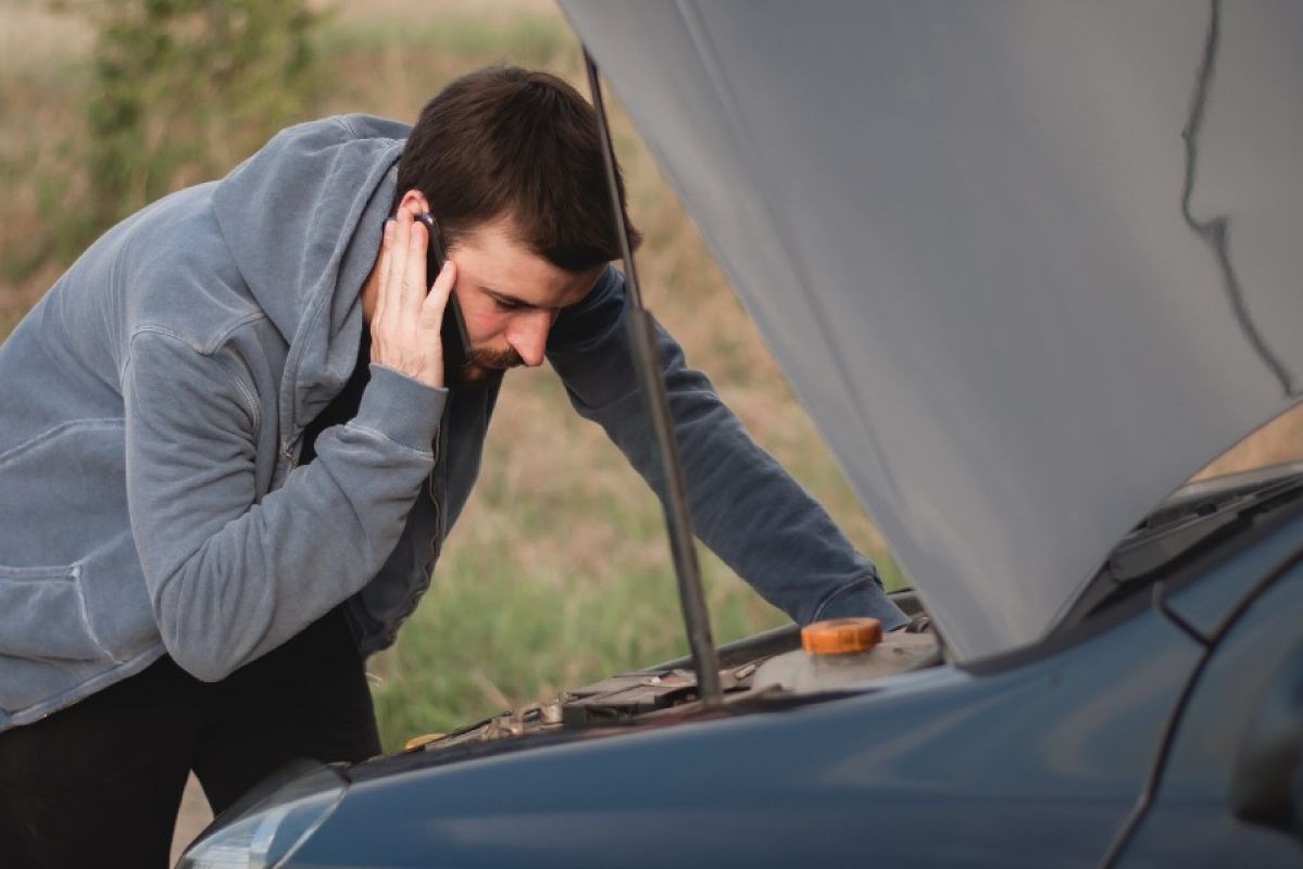 A man holding a phone up to his ear while he looks under the bonnet of his car, which is parked on the side of the road, near a field