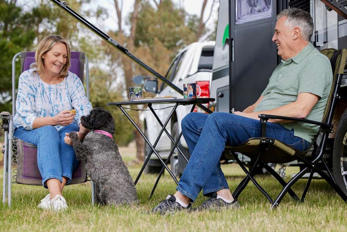 An older couple sitting on deck chairs in front of their parked caravan with their black spoodle dog