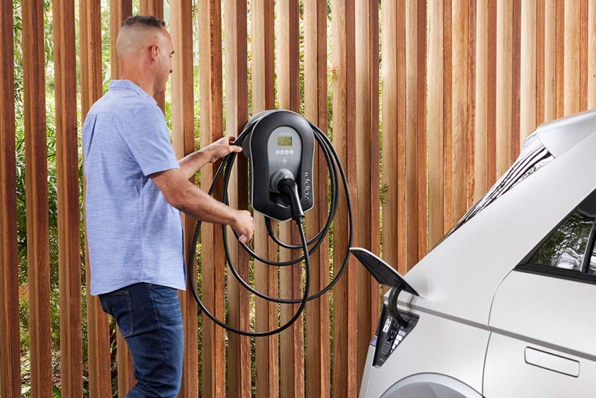 Man in blue shirt in carport getting connector from home EV charger on wall for white Hyundai Ioniq 5 EV in foreground
