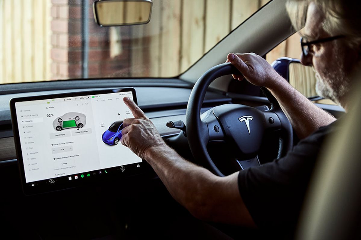Bearded man with glasses behind wheel of Tesla Model Y in carport checking EV charge status on touchscreen.