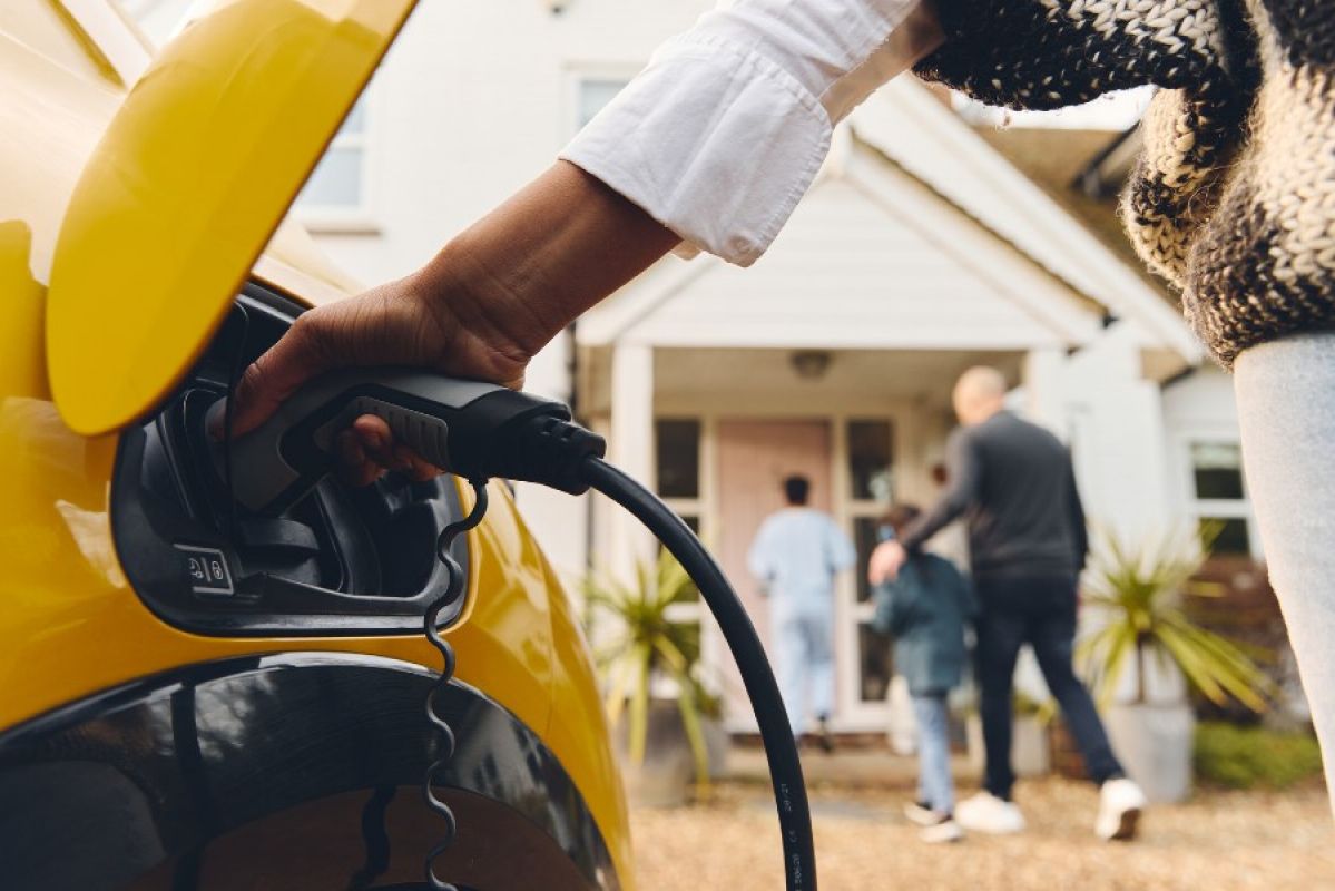 A close-up of a person putting a charging cable into their bright yellow EV. In the background a man and two children can ne seen walking towards a front door of a house