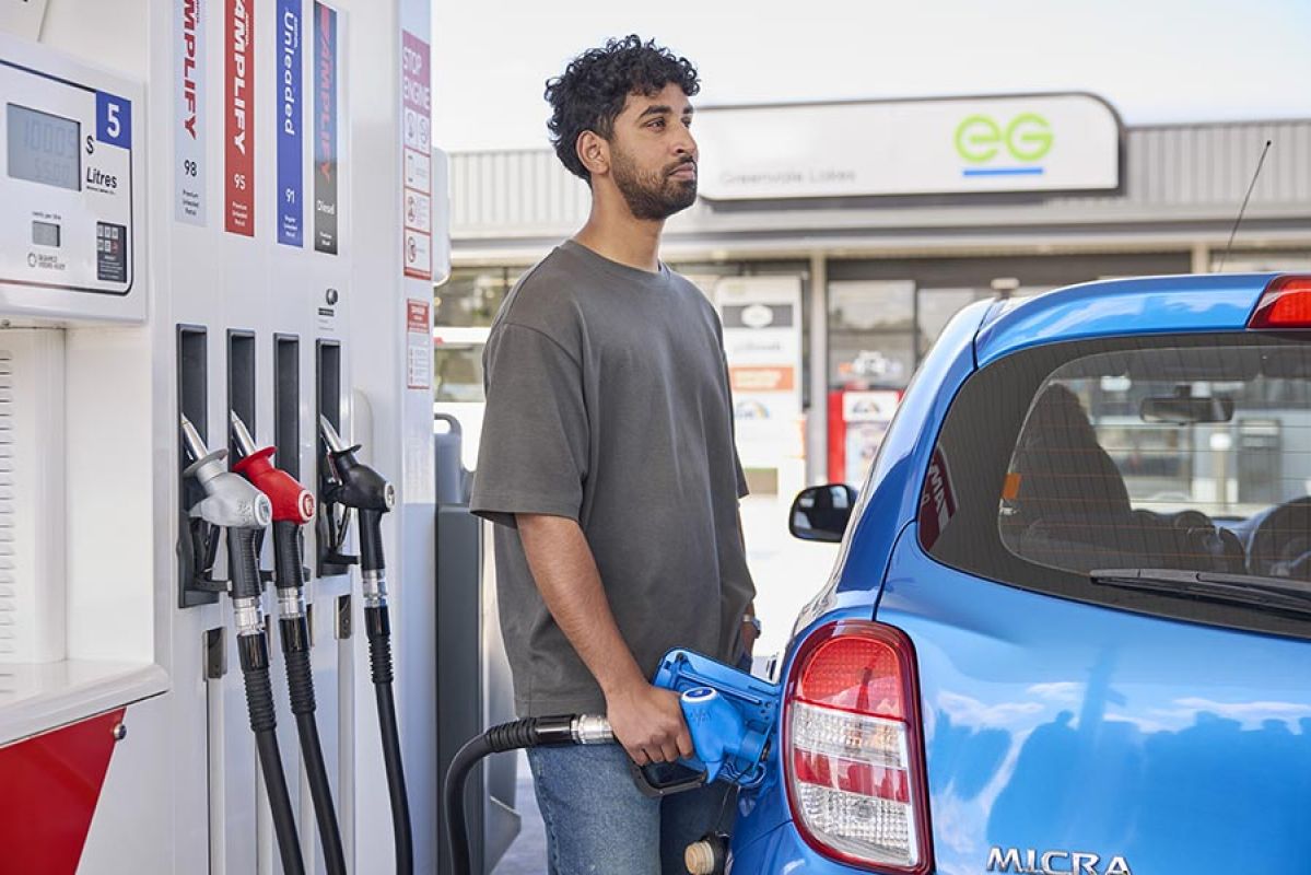 Bearded man with grey t-shirt looking into distance while filling up blue car at petrol station bowser.