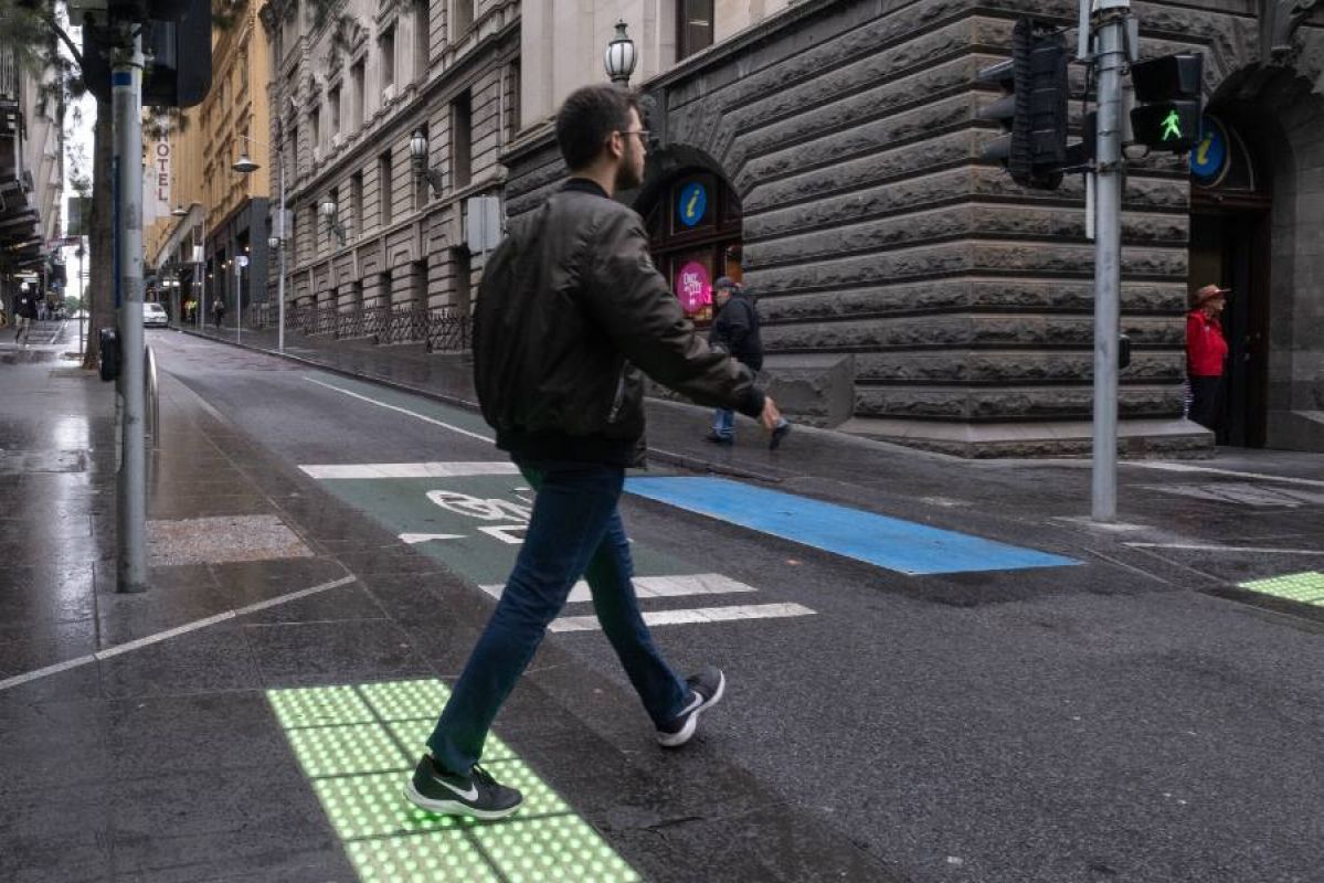 A man walking across a pedestrian crossing at the intersection of Swanston and Little Collins streets in Melbourne. It has been raining and the crossing features green light up tactile markers 