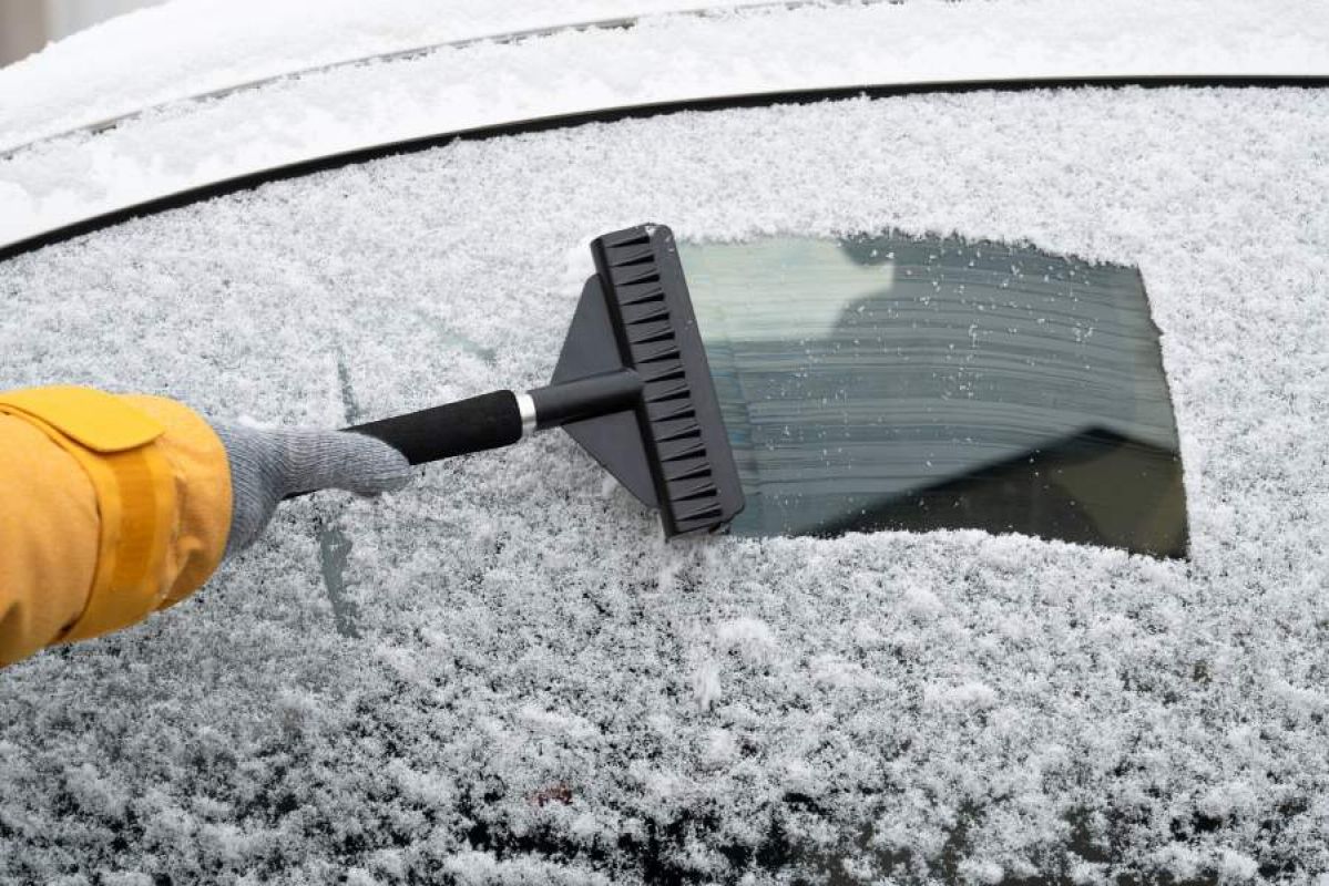 A person wearing a bright yellow snow jacket and gloves using a long-handled plastic  ice scraper to remove frost from their car windows