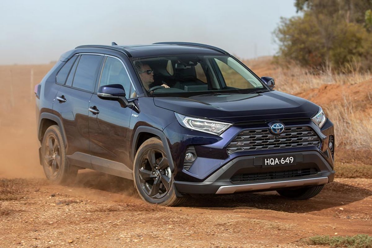 Man with sunglasses driving black Toyota RAV4 Hybrid SUV on dusty red dirt road next to dry agricultural land.