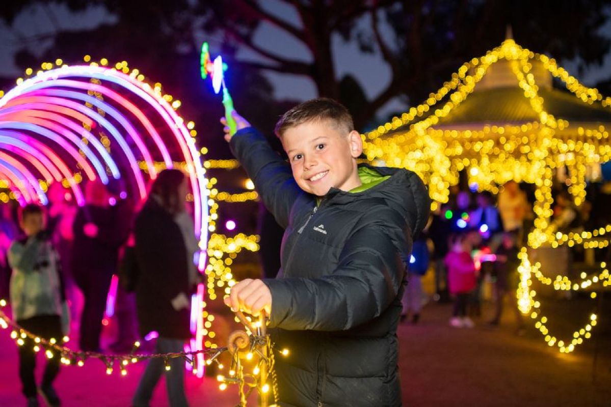A young boy smiles and holds onto a fairy light-lit fence post during Sovereign Hill's Winter Wonderlights festival. A tunnel of multicoloured lights and a gazebo lit by fairy lights can be seen in the background