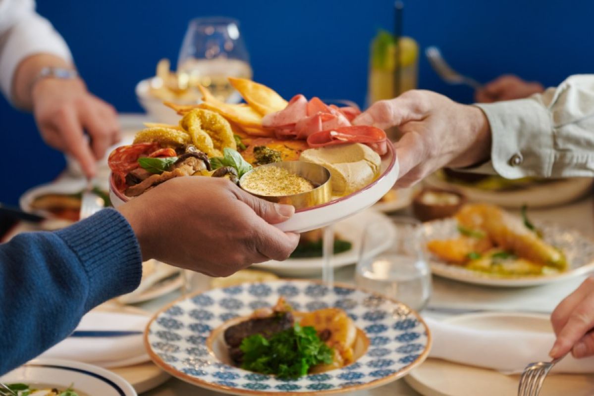 Two people passing a Greek-inspired mezze plate at Sojourn restaurant as part of a larger group meal