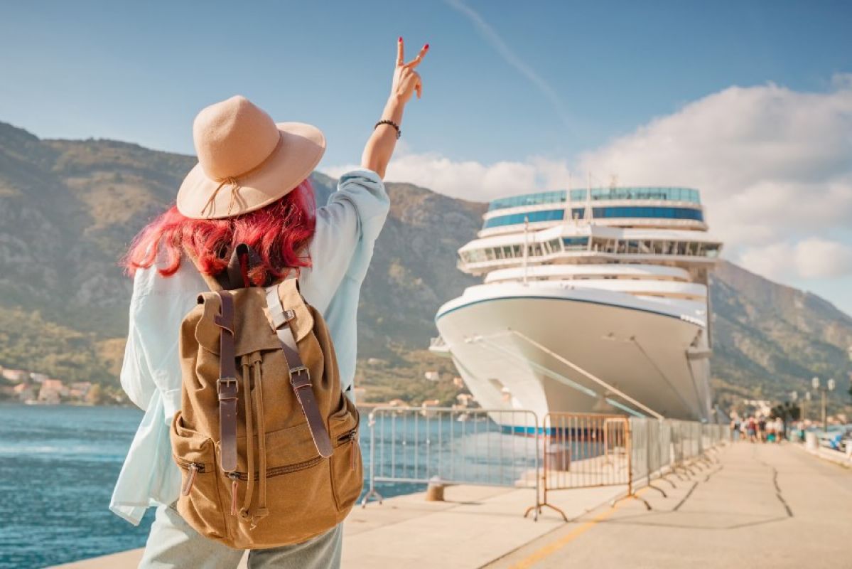 woman with red hair, sunhat and backpack giving a peace sign to a large cruise ship
