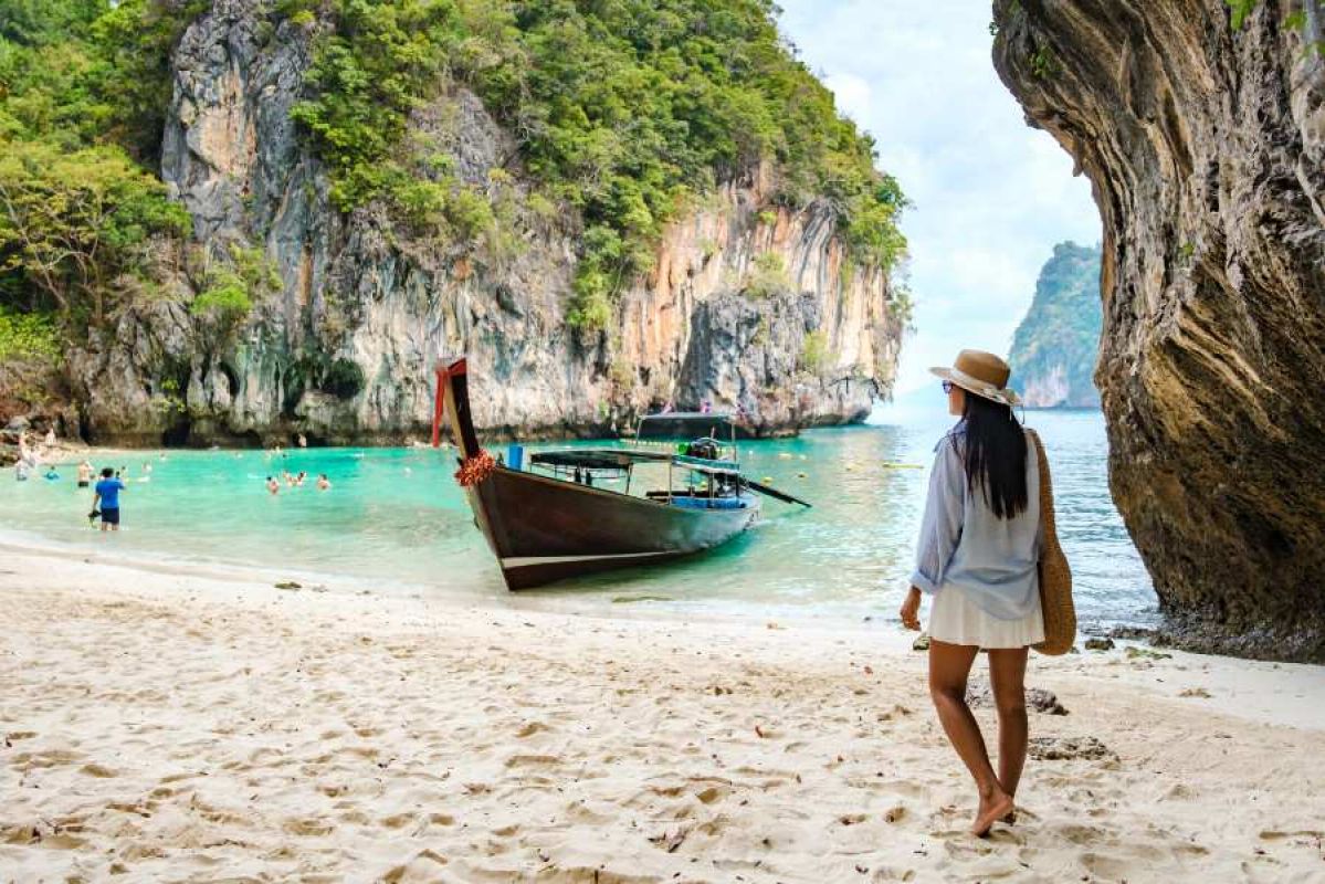 woman walking on a sandy Thai beach with boats, swimmers and islands in background