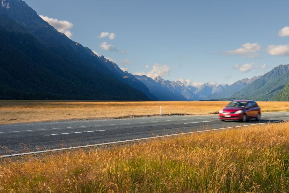 A small red hatchback car driving on a road approaching Milford Sound in New Zealand. The road is surrounded by golden plains of grass while tall mountains stand in the background