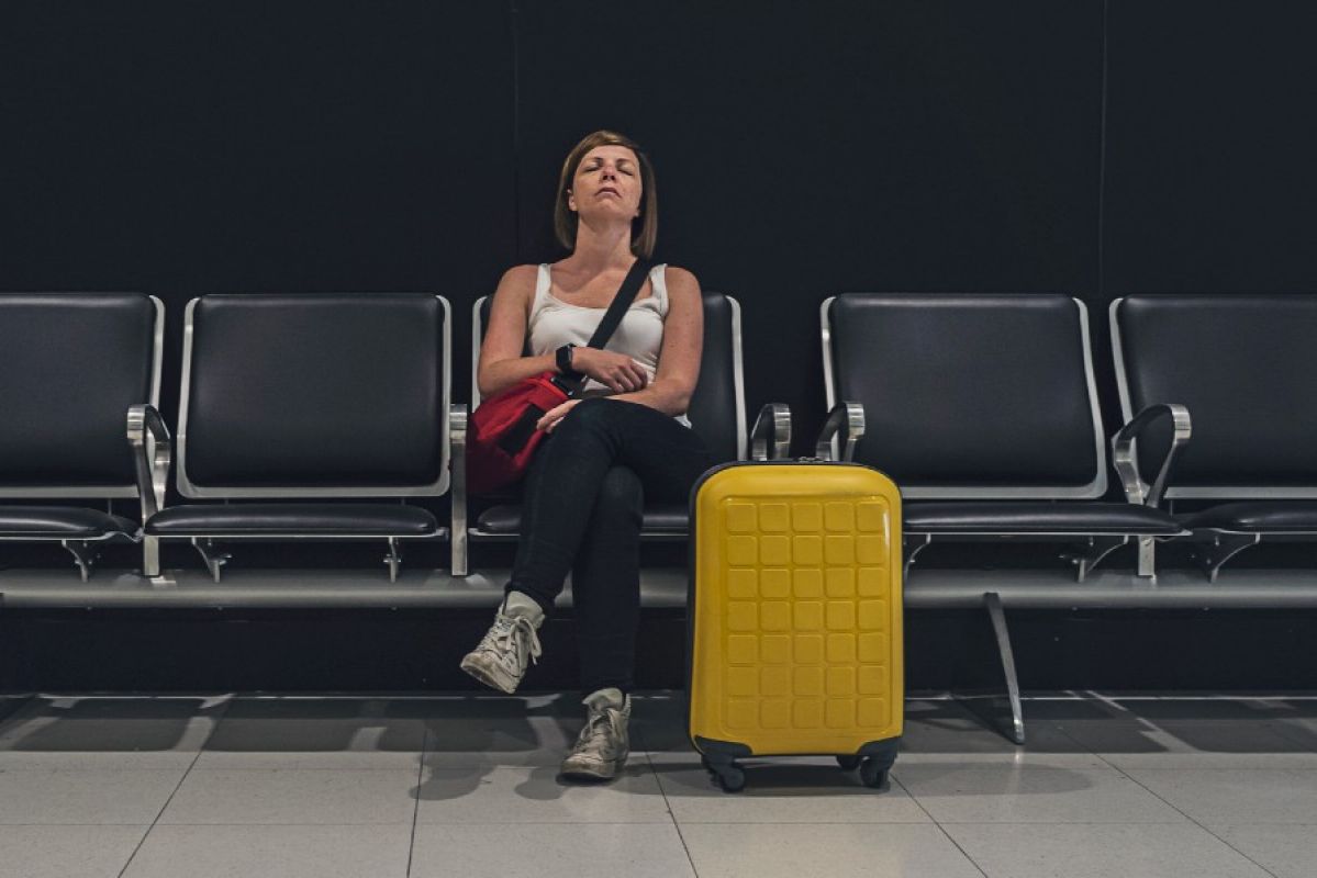 A tired-looking woman sitting on a row of seats waiting for her flight at an airport. She leans back on her chair with her eyes closed and legs crossed while a bright yellow suitcase is on the ground next to her