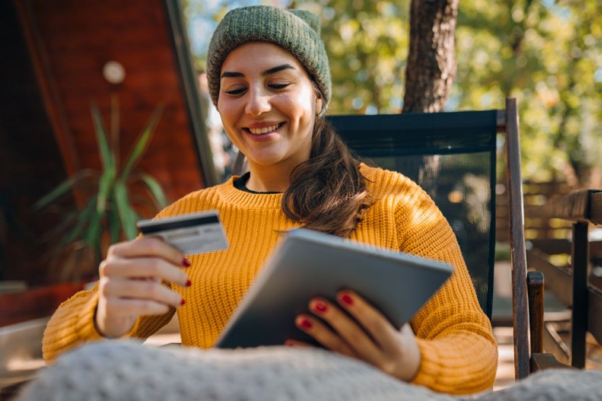 young woman in yellow sweater and green beanie sitting on a deck smiling at a credit card, holding a tablet
