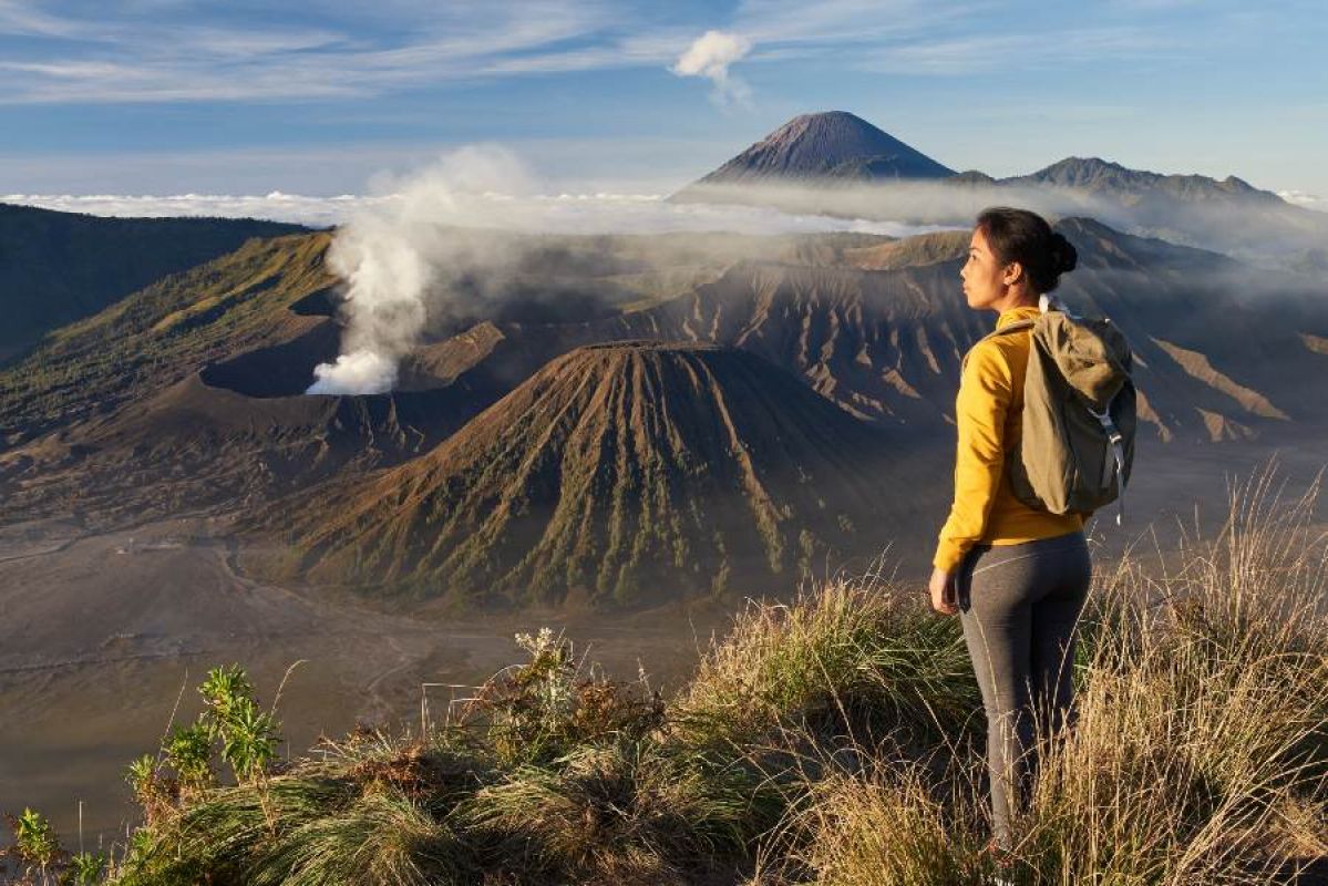 A woman wearing a yellow jacket, leggings and a backpack stands on a mountain overlooking a smoking Mount Bromo in Indonesia