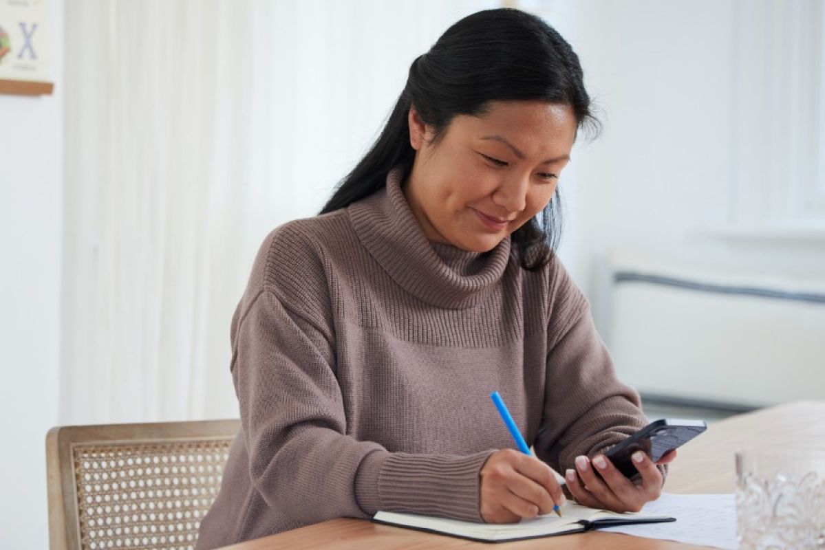 woman looking at smartphone and smiling while writing in a notepad