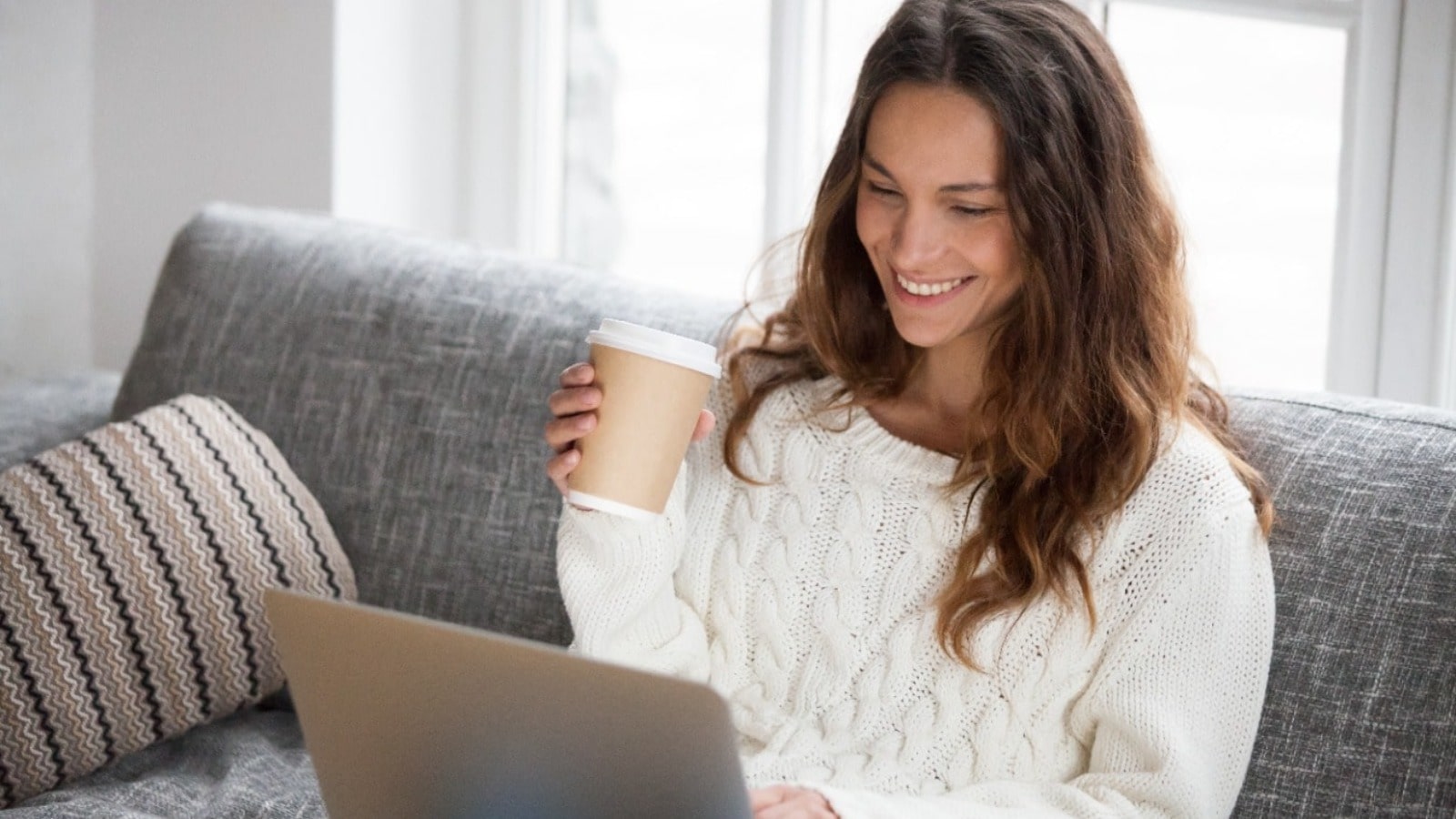 Woman sitting on couch, drinking takeaway coffee while looking at laptop.