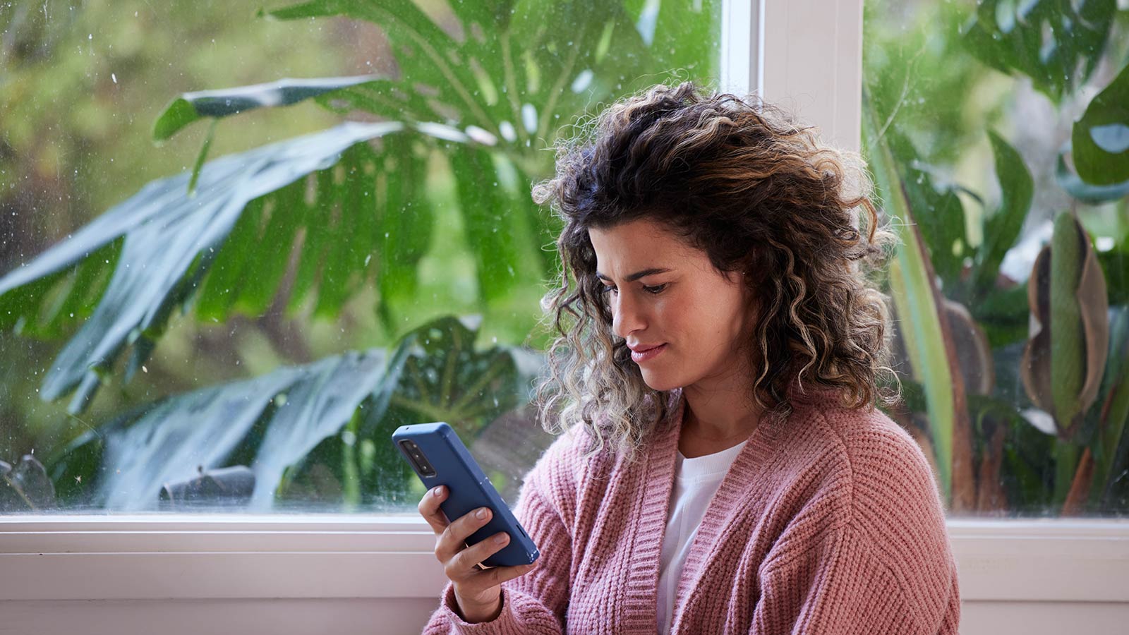 Woman with curly hair looking at her mobile phone while sitting in front of a window.
