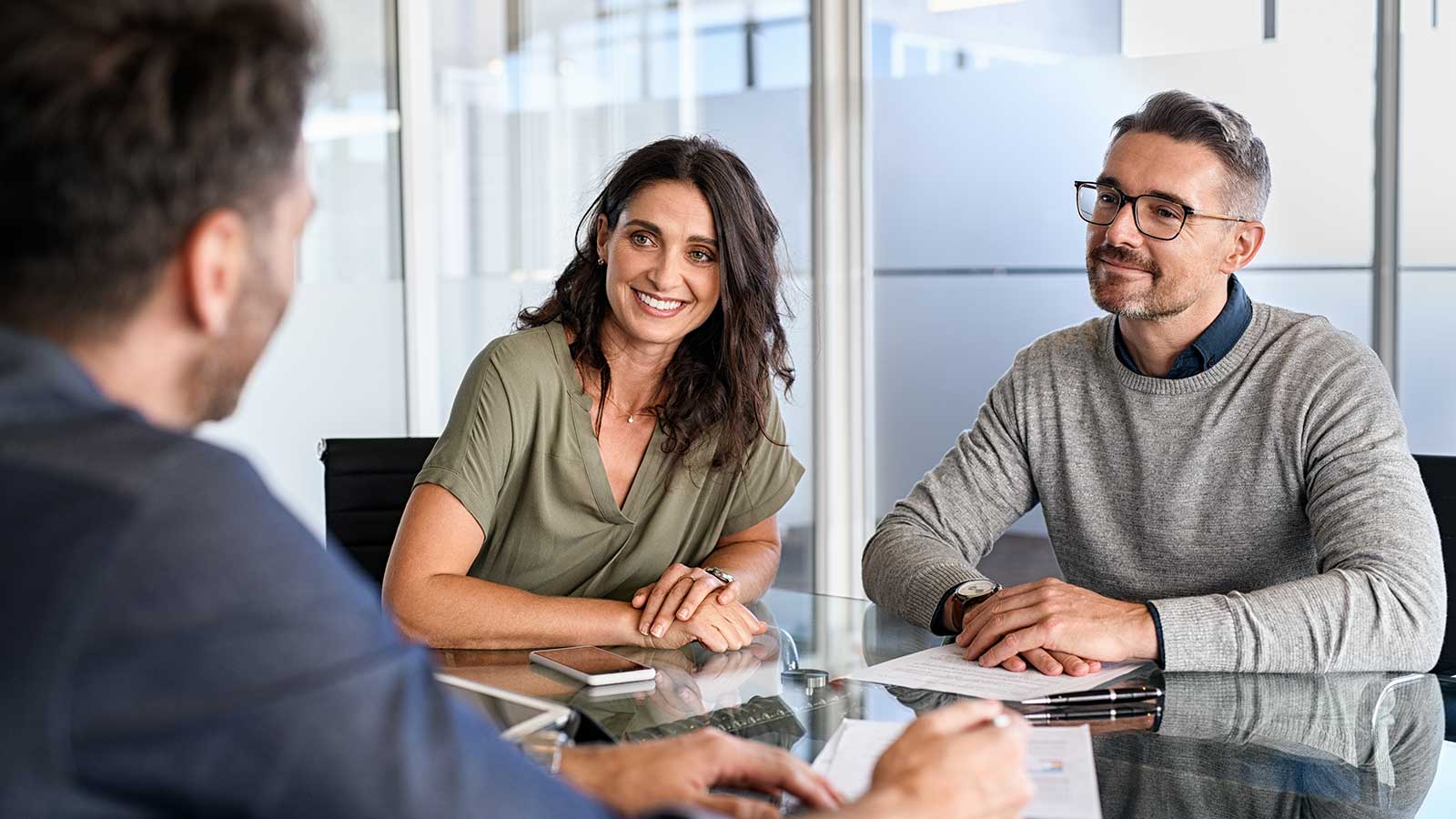 A smiling couple sit at a desk in front of a conveyancing lawyer who is preparing paperwork for signing. 