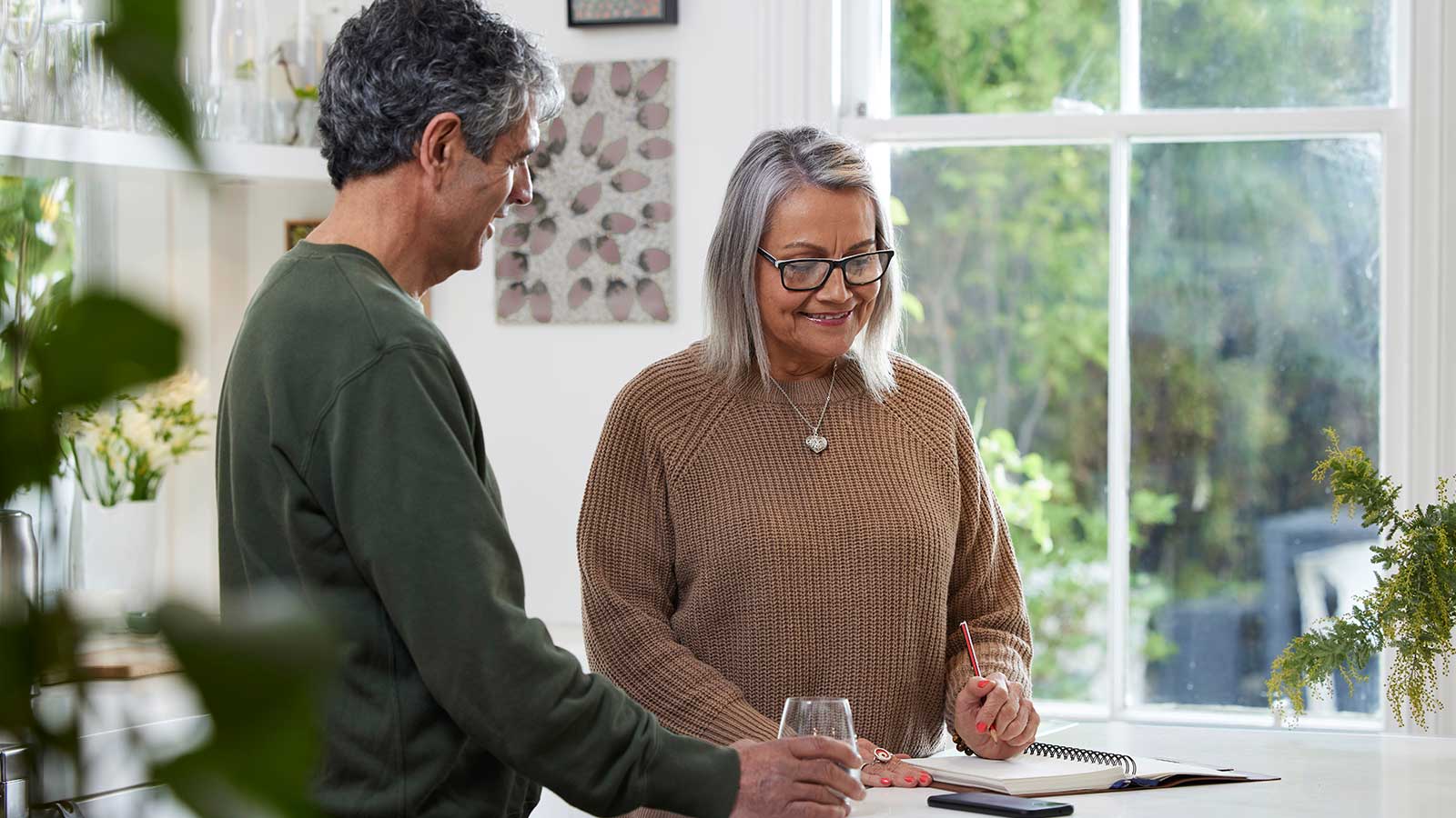 An older woman with grey hair and glasses stands in the kitchen in front of a bench, in front of her is an open notebook and a she is smiling. A smiling man with greying hair stands next to her, holding a glass of water, also smiling. 