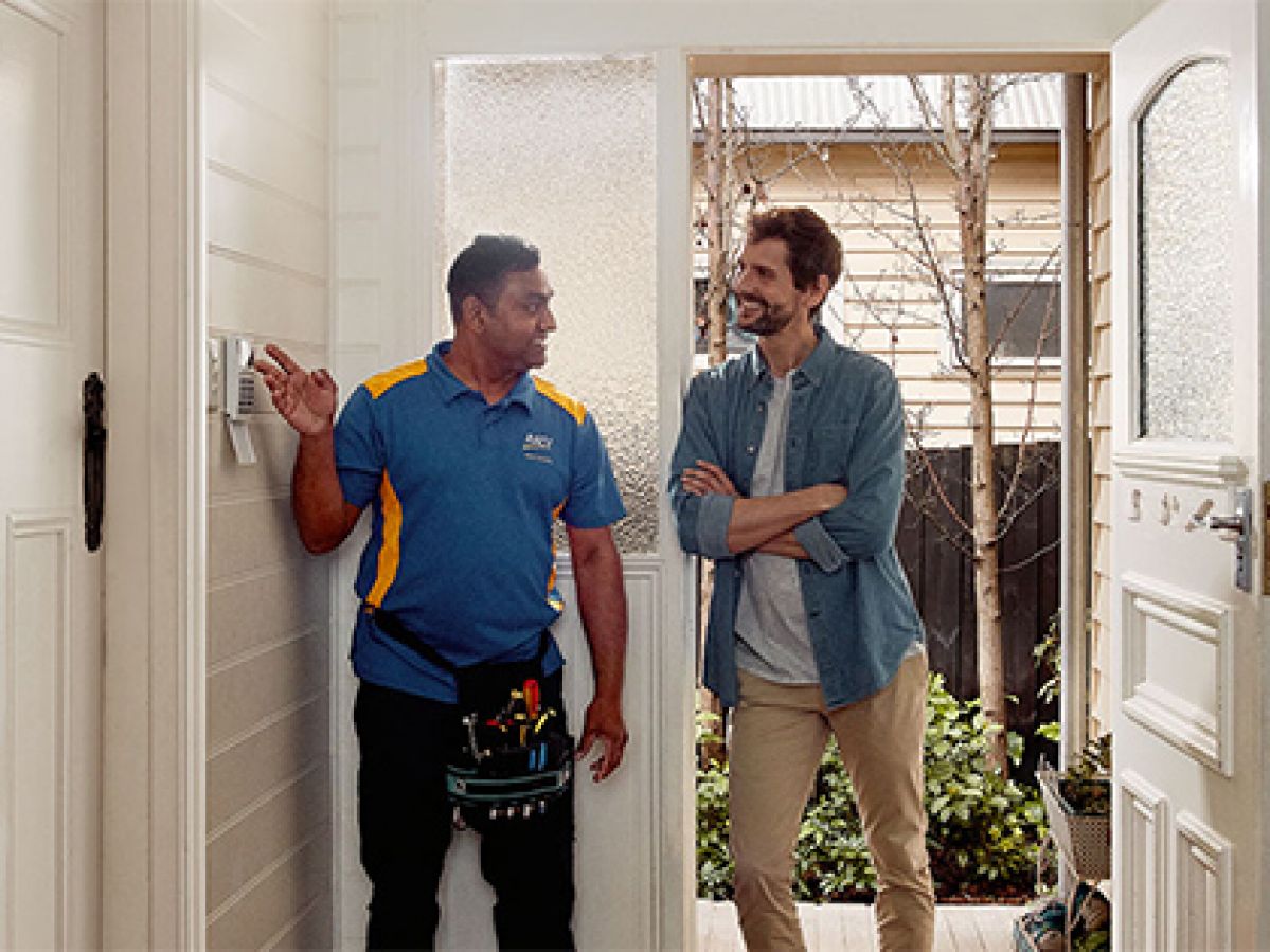 RACV Tradesman chatting to a member inside their house, whilst pointing at an alarm system.